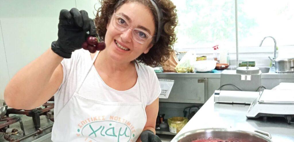 Μια γυναίκα με ποδιά στέκεται σε μια κουζίνα εργαστηρόιου και δείχνει κεράσια και μια κατσαρόλα που σιγοβράζει στο μάτι της κουζίνας με μαρμελάδα.