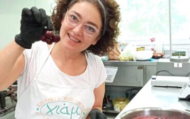 Μια γυναίκα με ποδιά στέκεται σε μια κουζίνα εργαστηρόιου και δείχνει κεράσια και μια κατσαρόλα που σιγοβράζει στο μάτι της κουζίνας με μαρμελάδα.